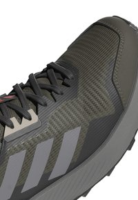 Baskets de sport vert foncé avec un tissu texturé, lacets noirs et trois rayures grises. Comprend un capuchon de protection renforcé et une semelle en caoutchouc.