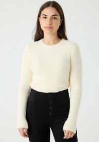 Mujer con cabello largo castaño, vistiendo un suéter blanco de manga larga y peludo, y pantalones negros de talle alto con cuatro botones frontales.