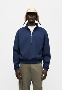 Μπλε ναυτικό φούτερ με φερμουάρ, φτιαγμένο από βαμβακερό μείγμα, με ριμπ μανσέτες και τελείωμα, σε συνδυασμό με μπεζ καπέλο τύπου bucket με επένδυση για τα αυτιά.