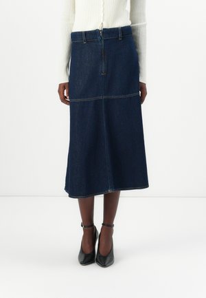 Dunkelblaue Denim-Midi-Rock mit tailliertem Bund, vorne mit Reißverschluss und sichtbaren Nähten. Kombiniert mit schwarzen Ankle-Strap-Absätzen.
