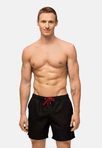 Pantaloni da nuoto neri con cordoncino rosso, tessuto leggero, vita elasticizzata e tasche laterali. Il modello mostra un fisico tonico.