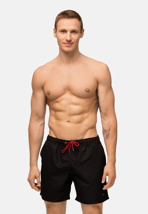 Bañador negro con cordón rojo, tejido ligero, cinturilla elástica y bolsillos laterales. El modelo muestra una figura tonificada.