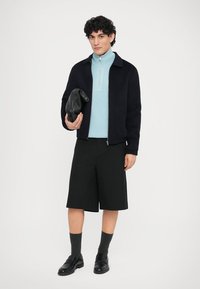Navy zip-up jacket, light blue high-neck sweater, black shorts, gray socks, black shoes, holding a black clutch bag. Simple design, modern cut.