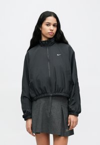 Μαύρο αθλητικό μπουφάν Nike με ψηλό γιακά, κοντό σχεδιασμό και ελαστική μέση. Διαθέτει λευκό λογότυπο στο αριστερό στήθος και φερμουάρ κλεισίματος.