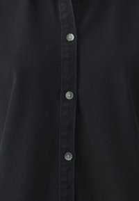 Camicia nera con bottoni, caratterizzata da una texture liscia, colletto classico e cinque bottoni di colore chiaro sul fronte.