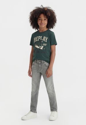 Dijete s kovrčavom kosom, odjeveno u tamnozeleno grafički printanu majicu, sive traperice i bijele tenisice, stoji na jednobojnoj pozadini.