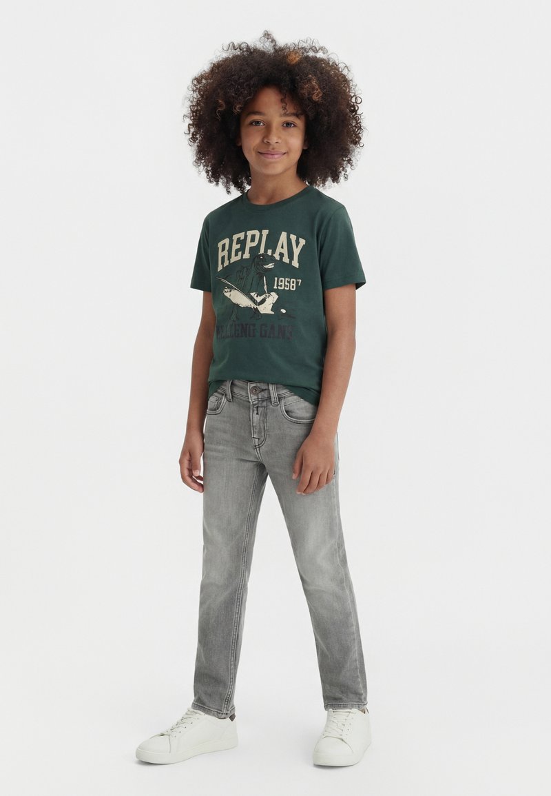 Παιδί με σγουρά μαλλιά που φοράει σκουροπράσινο μπλουζάκι με σχέδιο, γκρι τζιν και λευκά αθλητικά, στέκεται μπροστά σε απλό φόντο.