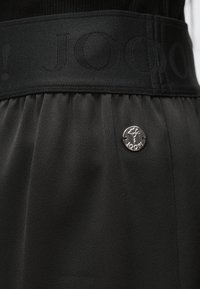 Fustă din material negru cu o bandă texturată în talie, având un logo în relief și un detaliu din capse argintii în față.