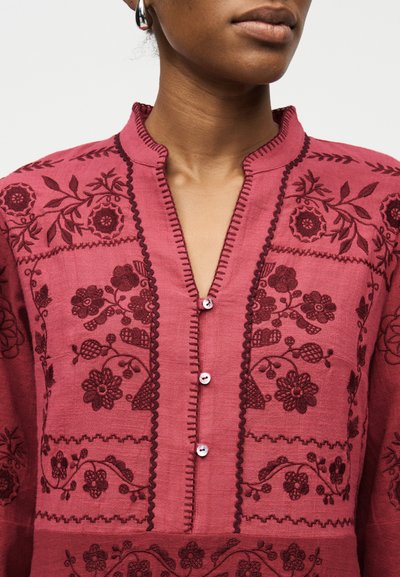 Γυναίκα που φοράει ένα βαθύ ροζ πουκάμισο με κουμπιά, σκοτεινή κεντητή λουλουδάτη διακόσμηση και γιακά μανταρίνι, εμφανιζόμενη από τον λαιμό μέχρι το μέσο του κορμού.