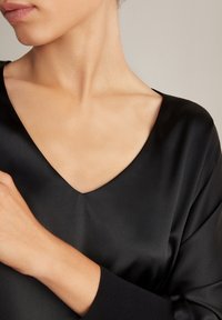 Top in satin nero con scollatura a V profonda e maniche lunghe. Il tessuto è liscio con una leggera lucentezza, evidenziando i contorni della scollatura.