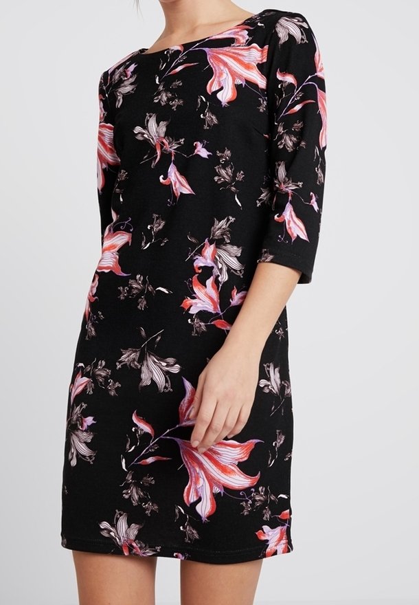 Femme portant une robe noire avec de grands motifs floraux roses et blancs et manches trois-quarts, debout devant un fond uni.