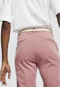 Pantalons rose clair avec une texture, dotés de poches arrière et d'une ceinture blanche tressée. La ceinture présente des détails de couture visibles.