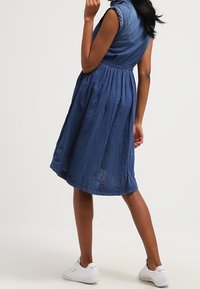 Sukienka jeansowa w głębokim niebieskim kolorze, z krótkimi rękawami, dopasowanym gorsetem i rozkloszowaną spódnicą. Noszona z białymi sneakersami, co podkreśla prosty, casualowy styl.