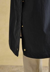 Jachetă ușoară neagră, cu nasturi aurii, textură netedă și hem ușor curbat, asociată cu pantaloni bej.