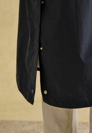 Schwarze leichte Jacke mit goldenen Druckknöpfen, einer glatten Textur und einem leicht abgerundeten Saum, kombiniert mit beigen Hosen.