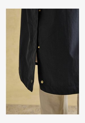 Čierna ľahká bunda s zlatými cvokmi, hladkou textúrou a jemne zakriveným lemom, v kombinácii s béžovými nohavicami.