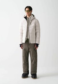 Giacca imbottita bianca leggera con cappuccio, dettagli con cerniere nere, abbinata a pantaloni cargo oliva con tasche e cerniere. Calzature nere.