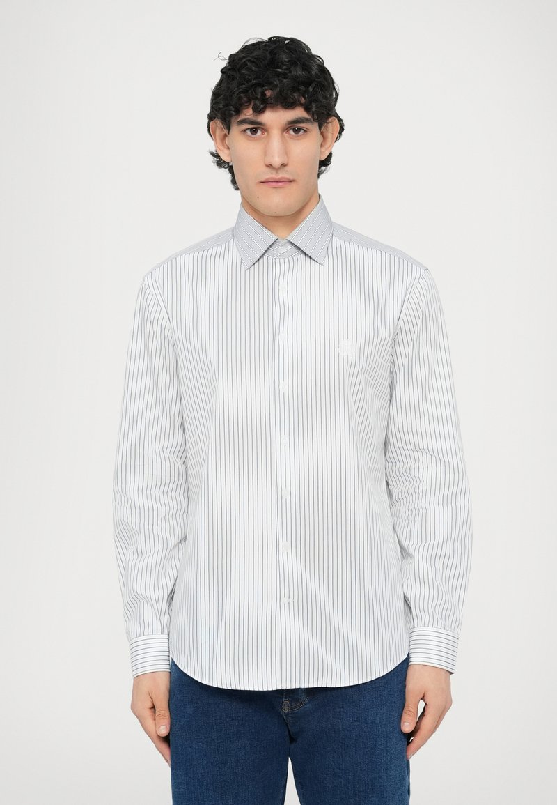 Biała koszula z długim rękawem w jasnoniebieskie pionowe paski, zapięty kołnierzyk na guziki i subtelne haftowane logo na piersi, w połączeniu z niebieskimi dżinsami.