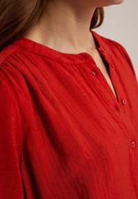 Red button-up shirt with a textured fabric. Features a shallow collar, five buttons, and gentle pleating near the shoulders.