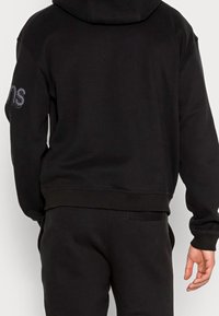 Persona con una sudadera negra con capucha y pantalones de chándal negros, mostrando la parte trasera con un texto reflectante parcial en la manga derecha.