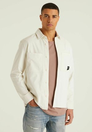White button-up shirt with a textured finish, featuring two front pockets and a logo patch, worn over a light pink t-shirt. Light blue distressed jeans.
