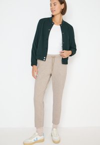 Tmavě zelená bomber bunda s knoflíky, bílý tričko s kulatým výstřihem, béžové vzorované tepláky a bílé tenisky s modrými akcenty.