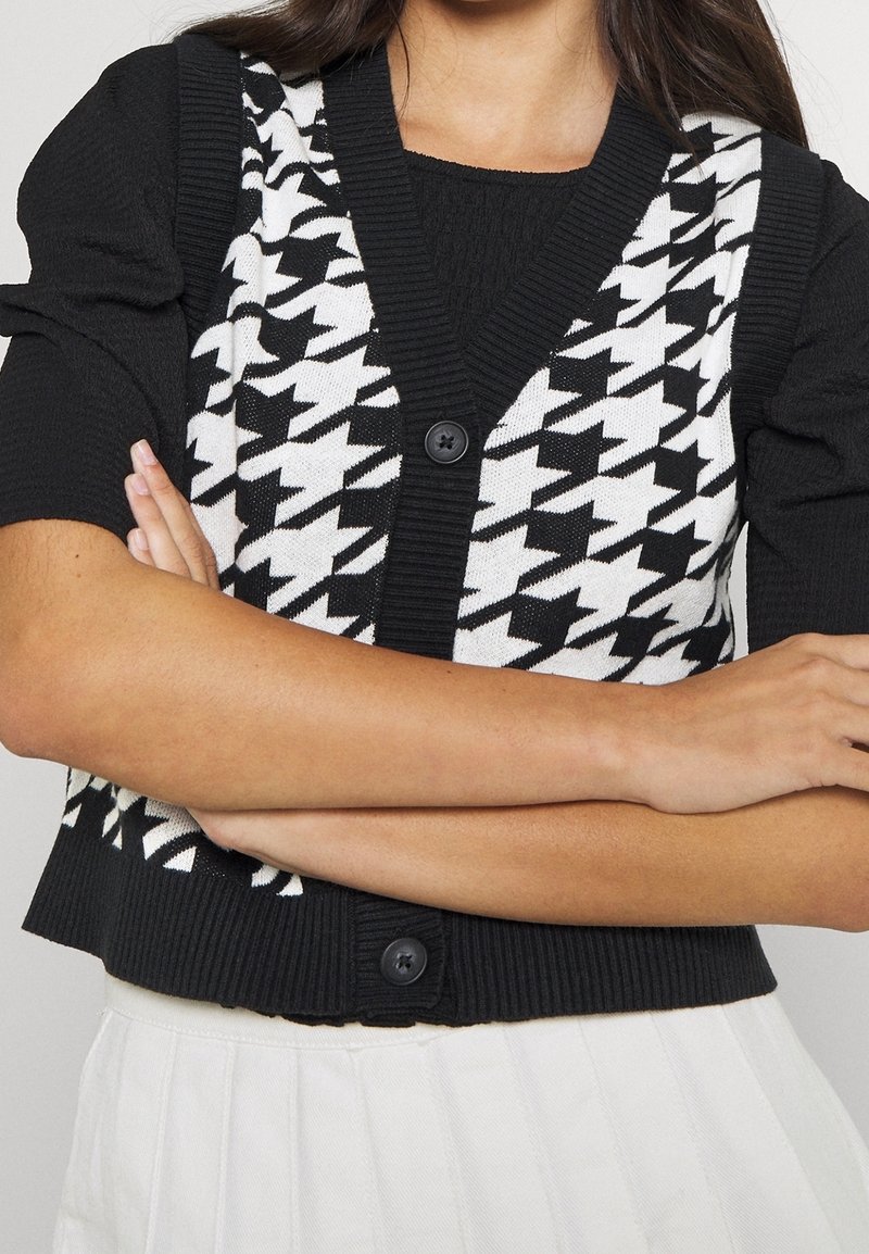 Personne croisant les bras, portant un haut noir à manches courtes avec un gilet boutonné à motif pied-de-poule noir et blanc, et une jupe blanche.