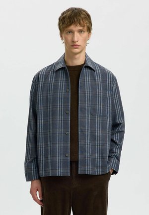 Jeune homme aux cheveux courts et bruns portant une chemise à carreaux bleue et grise boutonnée sur un pull marron foncé et un pantalon marron, debout face à l'avant.