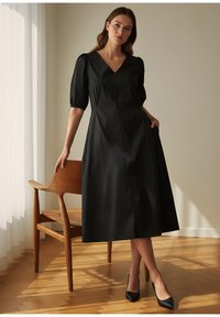 Črna midi obleka z V-izrezom in napihnjenimi rokavi. Gladek material, oprijet korzet, razširjen krilo. Model stoji ob lesenem stolu.
