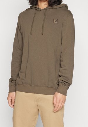 Personne portant un sweat à capuche vert olive avec cordons et un petit logo sur la poitrine, associé à un pantalon beige clair.