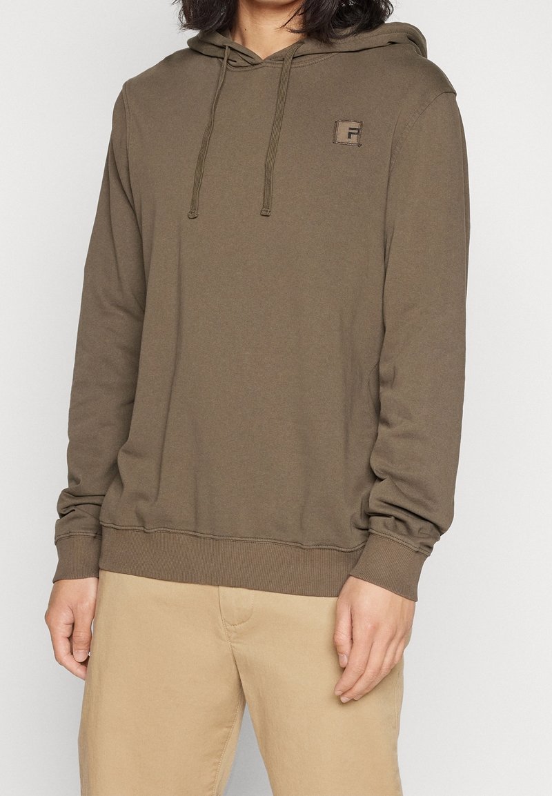 Personne portant un sweat à capuche vert olive avec cordons et un petit logo sur la poitrine, associé à un pantalon beige clair.