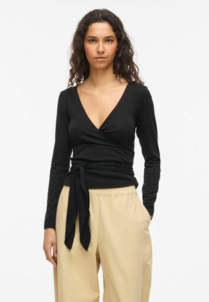 Žena nosí čierny dlhý rukáv zavinovací top so zaväzovaním v páse a béžové voľné nohavice, stojí na jednofarebnom bielom pozadí.