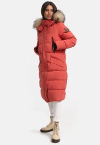 Palton puf roșu cu glugă căptușită cu blană, având fermoar frontal, capse și buzunare laterale mari. Purtați peste pantaloni albi și cizme bej.