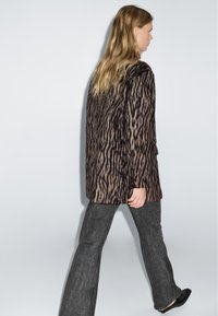 Cappotto in stampa animale marrone e crema con una texture morbida, caratterizzato da una vestibilità rilassata. Abbinato a pantaloni a zampa d'elefante grigi scuri e scarpe nere.