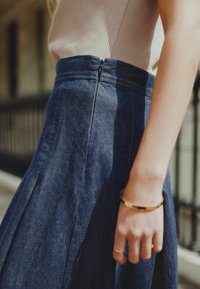 Denim sijonas su klostėmis, tamsiai mėlynos spalvos, su šoniniu užtrauktuku. Minimalistinis dizainas, prigludusi juosmens dalis ir sklandi tekstūra. Auksinė apyrankė kaip akcentas.