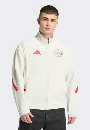 Ung mand iført en hvid lynlåstrøje med rødt Adidas-logo og grafisk emblem, kombineret med sorte jeans mod en ensfarvet baggrund.