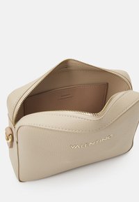 Beige leren tas met een gestructureerd oppervlak, gouden rits en een gelabeld binnenvak. Heeft een eenvoudige, rechthoekige vorm.