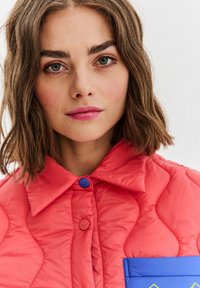 Mladá žena s krátkými hnědými vlasy v korálově prošívané bundě s modrým kapsou a růžovou rtěnkou na bílém pozadí.