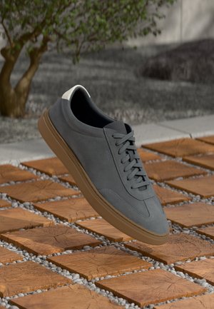 Gray suede sneaker with brown sole floating above wooden square tiles outdoors near a small tree and gravel ground.