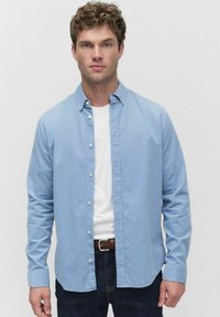 Lichtblauwe overhemdd met lange mouwen, gemaakt van gladde stof, gedragen over een wit T-shirt. Donkerblauwe jeans en een gevlochten riem maken de look compleet.