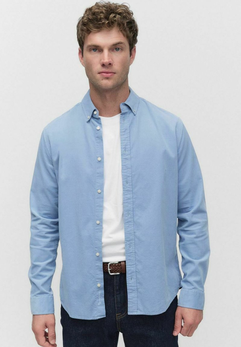 Lichtblauwe overhemdd met lange mouwen, gemaakt van gladde stof, gedragen over een wit T-shirt. Donkerblauwe jeans en een gevlochten riem maken de look compleet.