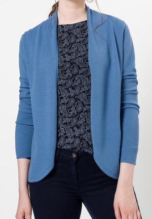 Cardigan texturé bleu à ouverture devant et manches trois-quarts, porté par-dessus un haut en motif paisley noir. Associé à un pantalon sombre.