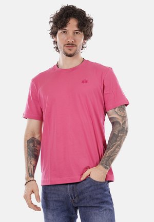 Uomo con capelli ricci che indossa una maglietta rosa e jeans blu, mostra tatuaggi dettagliati su entrambi gli avambracci, in piedi con una mano in tasca.