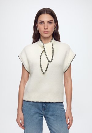 Vrouw met lang bruin haar draagt een witte mouwloze trui met zwarte stikselrand en lichtblauwe jeans tegen een effen witte achtergrond.