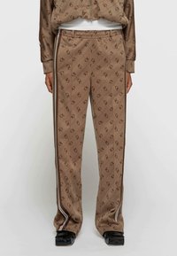 Brązowe wzorzyste spodnie dresowe z miękkiego materiału, z diagonalnym nadrukiem logo, bocznymi paskami i luźnym krojem dla wygody.