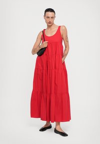 Abito maxiblu in cotone rosso con scollatura a barca, design a strati e orlo a balze. Indossato con una piccola borsa nera a tracolla e scarpe nere flat.