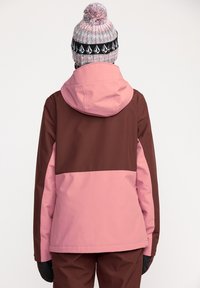 Chaqueta de esquí rosa y marrón con capucha y puños elásticos, con un diseño de bloques de color. Gorro de punto en rosa y gris con banda negra y patrones.