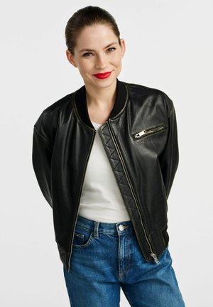 Czarna skórzana kurtka bombowiec o pikowanej fakturze, z ribbowanym kołnierzem i zamkniętą kieszenią na piersi, zestawiona z białą koszulą i niebieskimi dżinsami.