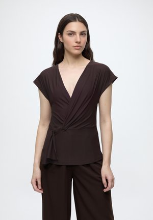 Femme aux longs cheveux foncés portant un top cache-cœur marron foncé à manches courtes et un pantalon à jambes larges assorti, sur un fond uni.