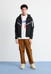 Casaco preto com fecho zip, com detalhes refletivos, sobre uma t-shirt gráfica branca, combinado com calças castanhas e ténis bege com cadarços vermelhos.